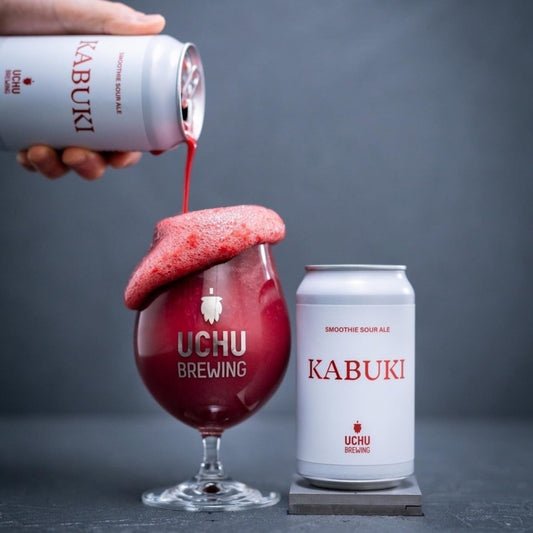 Kabuki beer being poured into a Uchu Brewing glass with two cans of Kabuki beer on a gray background.